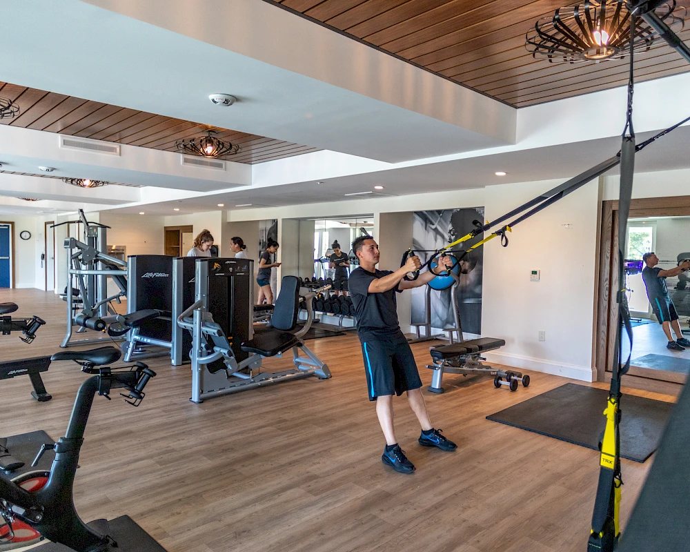 Someone working out in a well-equipped gym, using a suspension trainer and cable machines, while others exercise in the background.