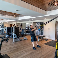 Someone working out in a well-equipped gym, using a suspension trainer and cable machines, while others exercise in the background.