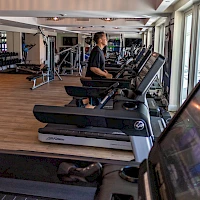 In a bright gym, a man runs on a treadmill while others and various cardio machines line a spacious, well-lit workout floor.