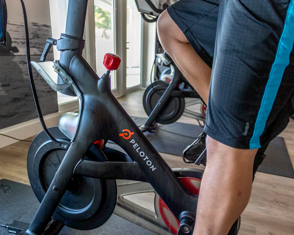 A person is riding a black Peloton stationary bike indoors, pedaling vigorously with a window-lit gym setting in the background.