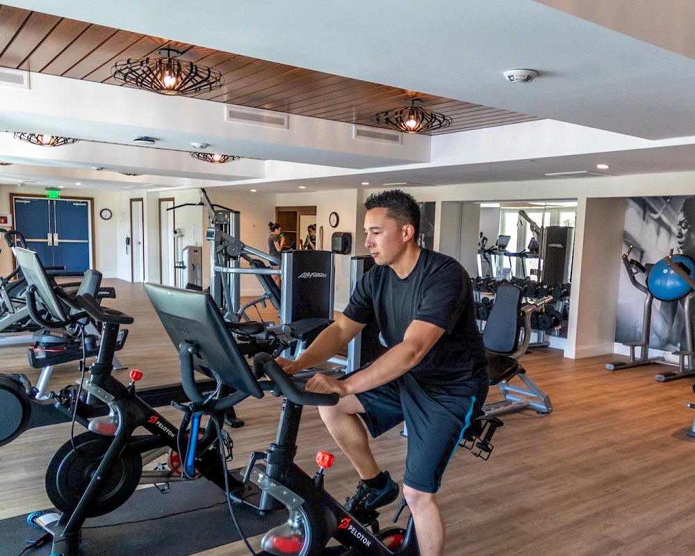 A man works out on a stationary bike in a bright gym with treadmills and other cardio machines, wooden ceiling panels, and mirrors, mid-workout.