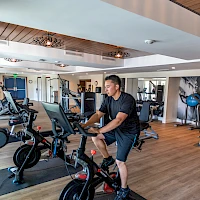 A man works out on a stationary bike in a bright gym with treadmills and other cardio machines, wooden ceiling panels, and mirrors, mid-workout.