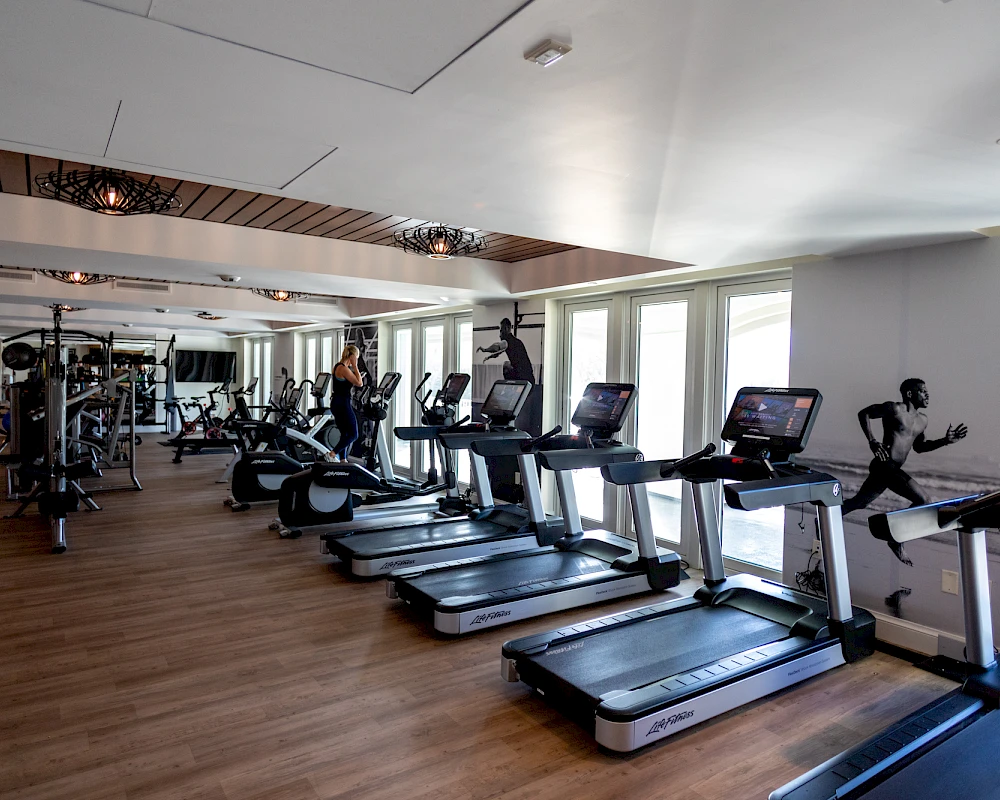 A spacious gym with multiple treadmills and people exercising, plus weight machines and mirrors along the back wall.