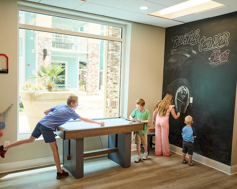 Kids playing air hockey and drawing on a chalk wall in a bright indoor playroom; a window shows a sunny courtyard outside.