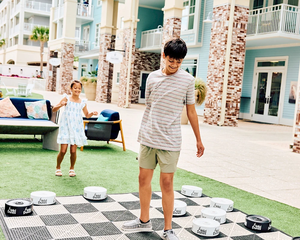 Two kids play life-sized chess on a board outdoors, with houses and seating in the background, enjoying a sunny scene.