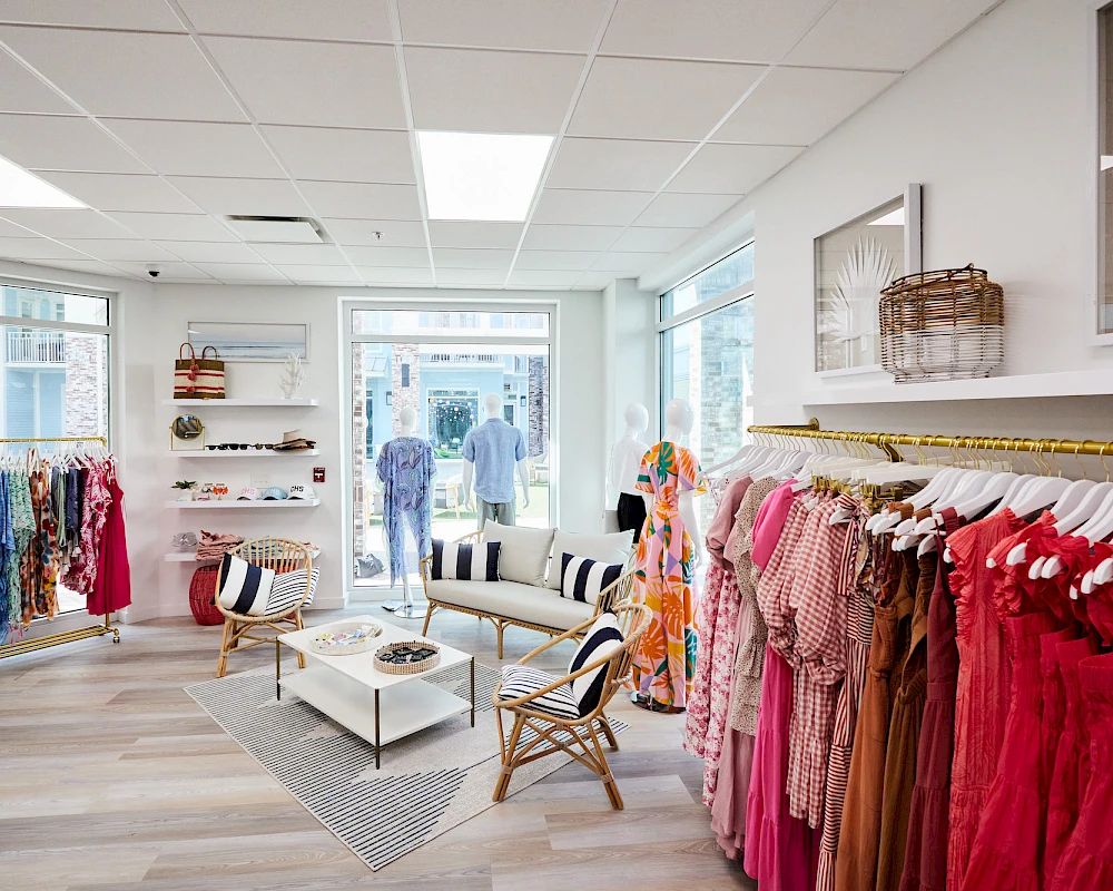 A bright boutique with racks of colorful clothes, stylish seating, a patterned rug, and large front windows letting in natural light.