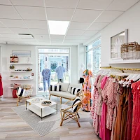 A bright boutique with racks of colorful clothes, stylish seating, a patterned rug, and large front windows letting in natural light.