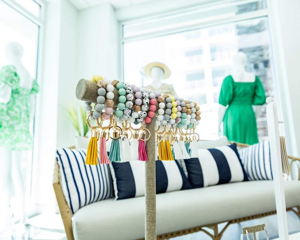 A bright boutique with a striped sofa, colorful beaded jewelry display, and green dress on a mannequin by a window, sunny and inviting.