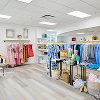 A bright boutique with light wood floors, racks of colorful dresses and tops, shelves of accessories, and a cozy seating area for shoppers.