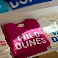 T-shirts on a rack with "WILD DUNES" in bold letters, white, pink, and cream colors, shop display at a clothing store.