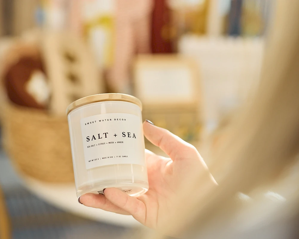 A hand holds a white candle labeled “Salt + Sea” in a cozy shop, soft focus in the background.