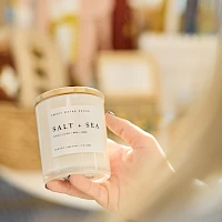 A hand holds a white candle labeled “Salt + Sea” in a cozy shop, soft focus in the background.