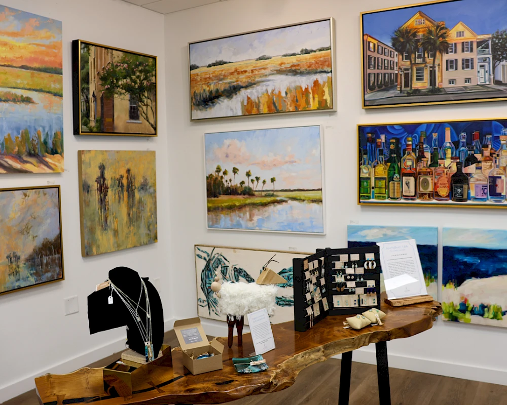 Colorful gallery wall with coastal and urban paintings, plus wooden sculptures and jewelry on a table.