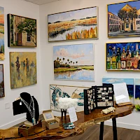 Colorful gallery wall with coastal and urban paintings, plus wooden sculptures and jewelry on a table.
