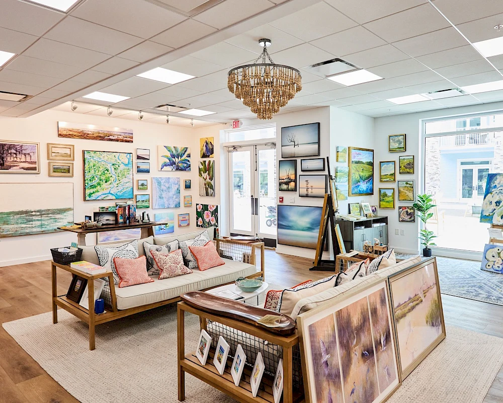A bright art gallery or boutique with paintings on the walls, a chandelier, wooden sofas, and art easels displaying colorful canvases.