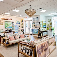 A bright art gallery or boutique with paintings on the walls, a chandelier, wooden sofas, and art easels displaying colorful canvases.