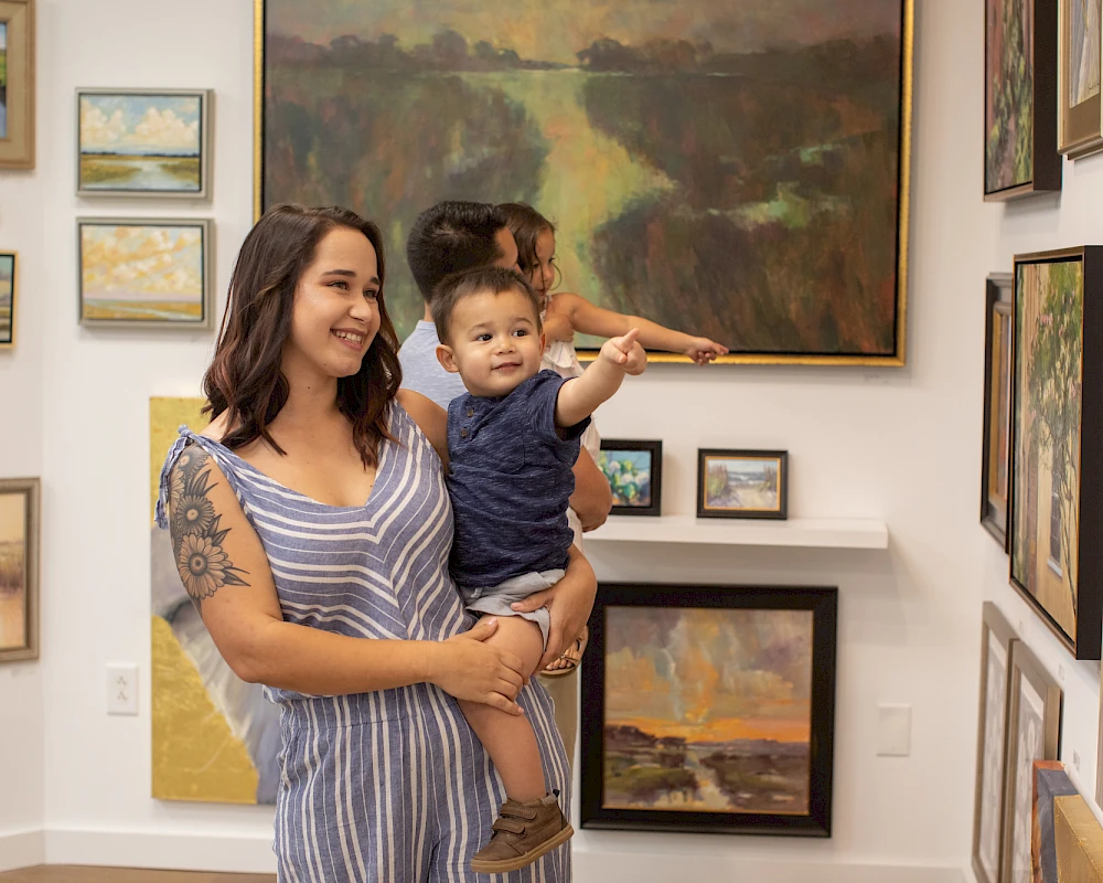 A smiling woman holds a toddler in a bright art gallery, surrounded by framed landscape paintings on white walls.
