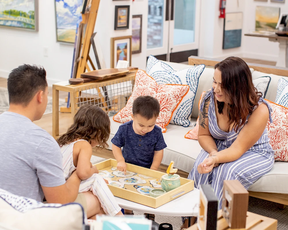 A family sits in a cozy art-filled living room, kids looking at books on a low table while parents chat, surrounded by framed artwork and cushions.