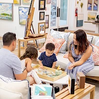 A family sits in a cozy art-filled living room, kids looking at books on a low table while parents chat, surrounded by framed artwork and cushions.