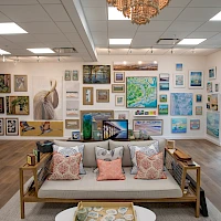 A cozy art gallery with a sofa, colorful cushions, and numerous paintings hung on white walls.