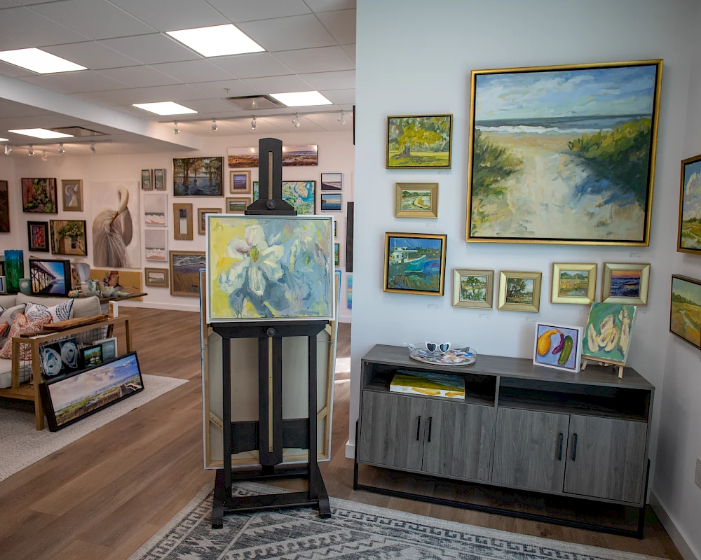 In a bright gallery, many paintings adorn walls and stands; a cabinet and easel hold colorful art, with a cozy seating area visible in the background.