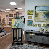 In a bright gallery, many paintings adorn walls and stands; a cabinet and easel hold colorful art, with a cozy seating area visible in the background.