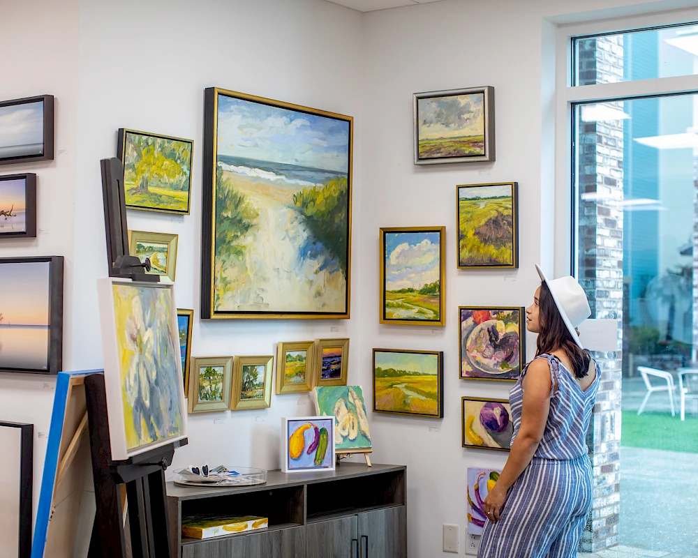 A woman in a striped dress and sun hat stands in an art gallery admiring a wall of landscape paintings arranged in various frames, with a windowed door to a bright outdoor area.