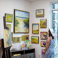 A woman in a striped dress and sun hat stands in an art gallery admiring a wall of landscape paintings arranged in various frames, with a windowed door to a bright outdoor area.