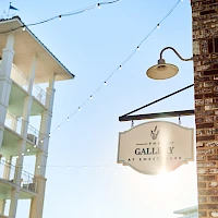 A sunny street scene featuring a brick building with a hanging lamp, string lights, and a sign that reads "Gallery" between pastel-colored apartment blocks.