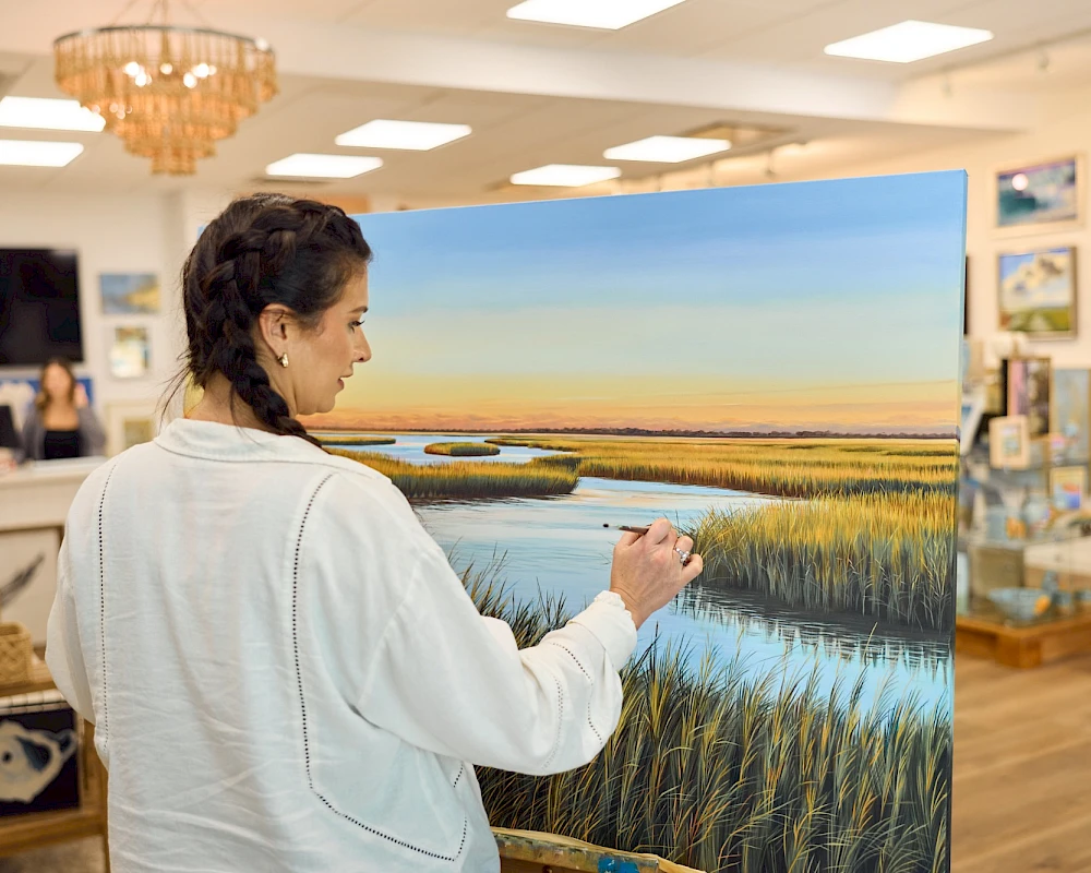 A woman in a light jacket paints a large landscape on a canvas in a bright gallery, featuring a sunset over marshland with tall grasses.