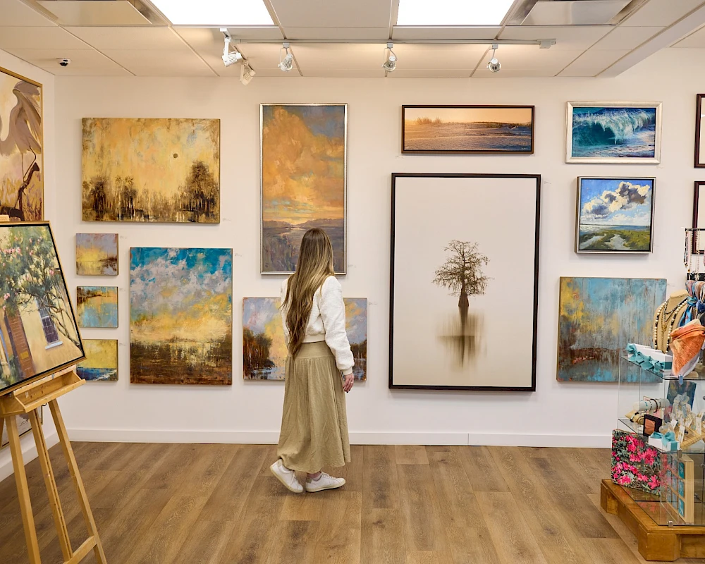 A person in a gallery viewing a variety of landscape and nature paintings on the walls and on display shelves.