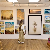 A person in a gallery viewing a variety of landscape and nature paintings on the walls and on display shelves.