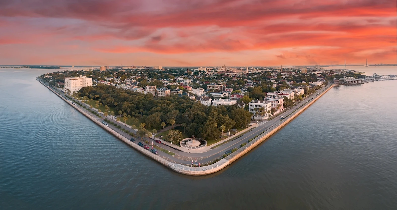 Aerial view of a triangular peninsula with a tree-filled park, surrounding water, and low-rise buildings; dramatic red sunset sky above.