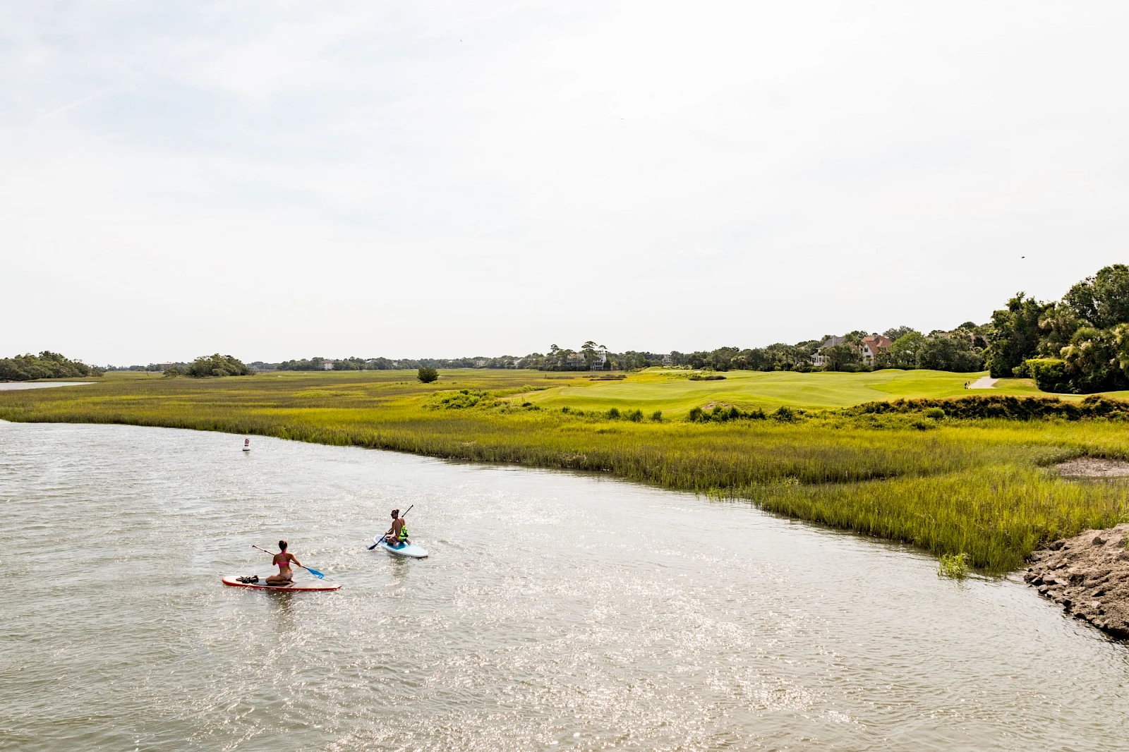 Two people kayak along a calm river estuary with grassy banks and distant trees under a bright, clouded sky, enjoying a peaceful paddle.