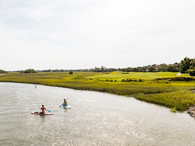 Two people kayak along a calm river estuary with grassy banks and distant trees under a bright, clouded sky, enjoying a peaceful paddle.