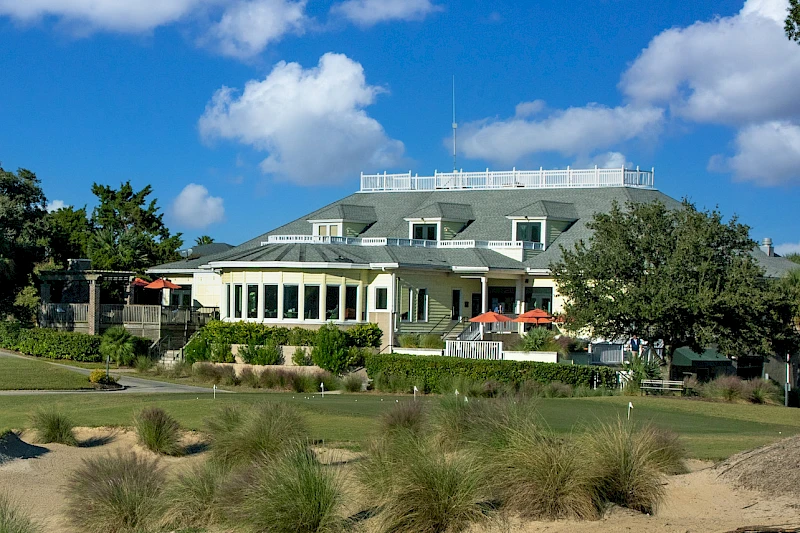 A large oceanfront clubhouse with a wraparound porch, manicured grounds, and a bright blue sky, shaded by trees and grasses.
