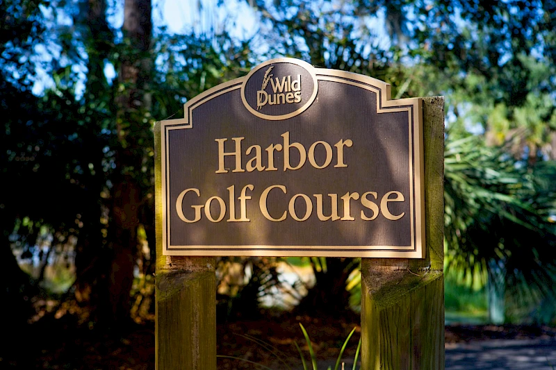 A sign for Harbor Golf Course at Wild Dunes, set among trees and lush greenery, inviting golfers to play on this sunny course.