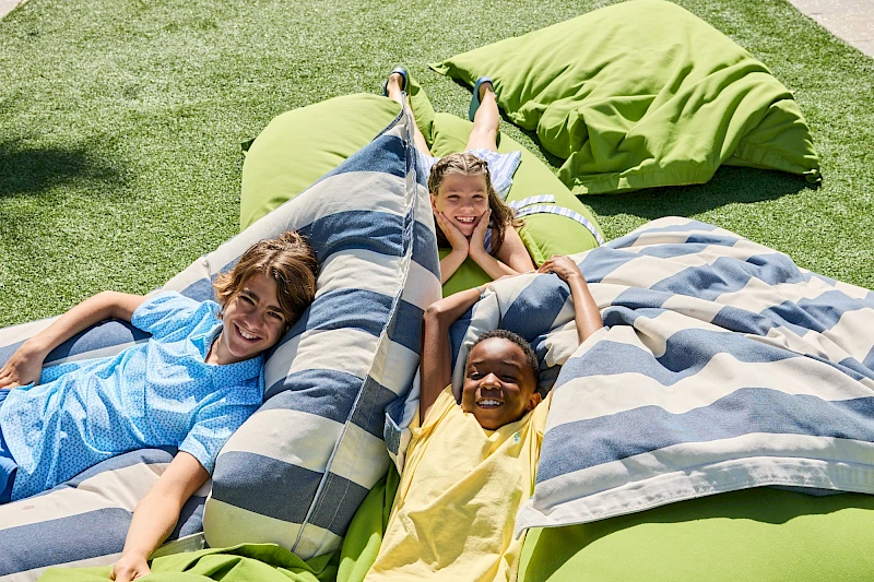 Three kids lounge on large striped and green beanbags outdoors, smiling and relaxing on a sunny grassy yard.