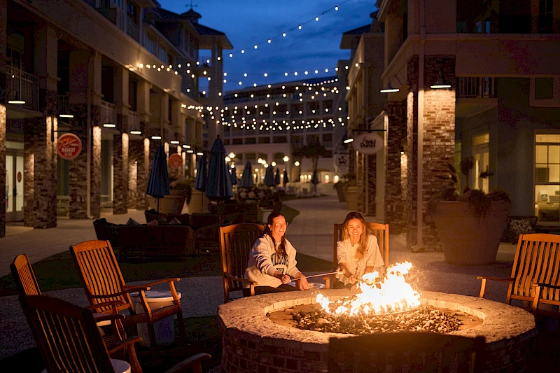 Two people sit by a glowing fire pit in a cozy outdoor village at dusk, string lights overhead, chairs around, nightlife ambiance, warm glow.