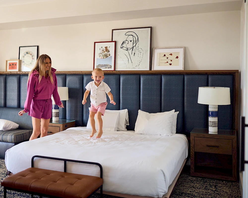 Two kids jump on a large hotel bed while a woman watches; stylish bedroom with framed art, blue upholstered headboard, and matching nightstands.
