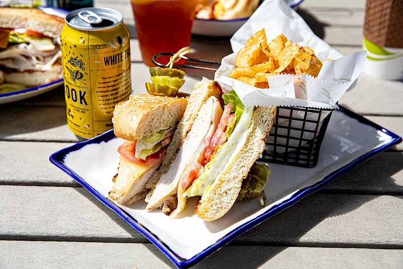 A plate with a club sandwich, potato chips in a basket, a yellow can of drink, and a glass of iced tea on a wooden table.