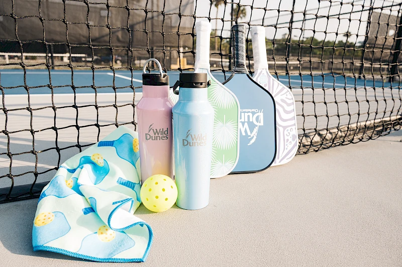 A tennis court scene with a net in the background; water bottles, a ball, a blue towel, and a couple of rackets cases or paddles lined up by the net.