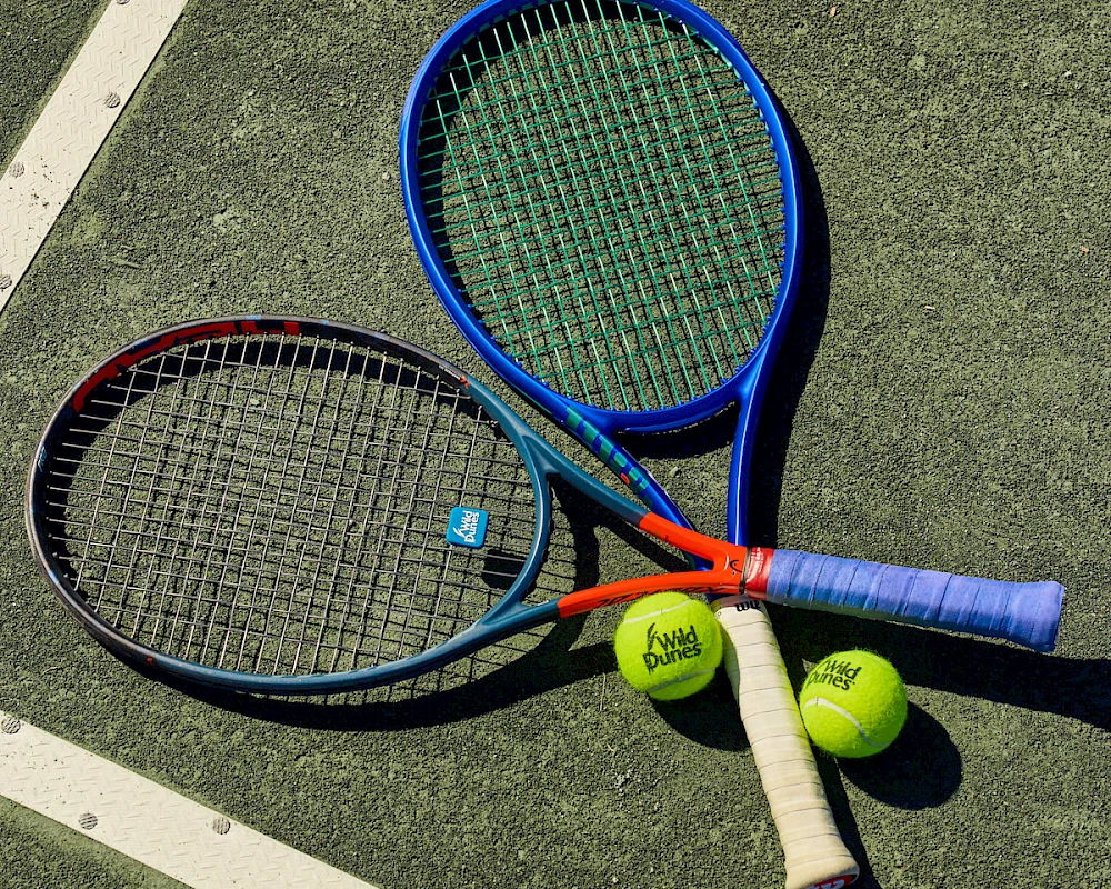 Two tennis rackets and three tennis balls on a sunlit court, ready for a match, with a white boundary line in the background.