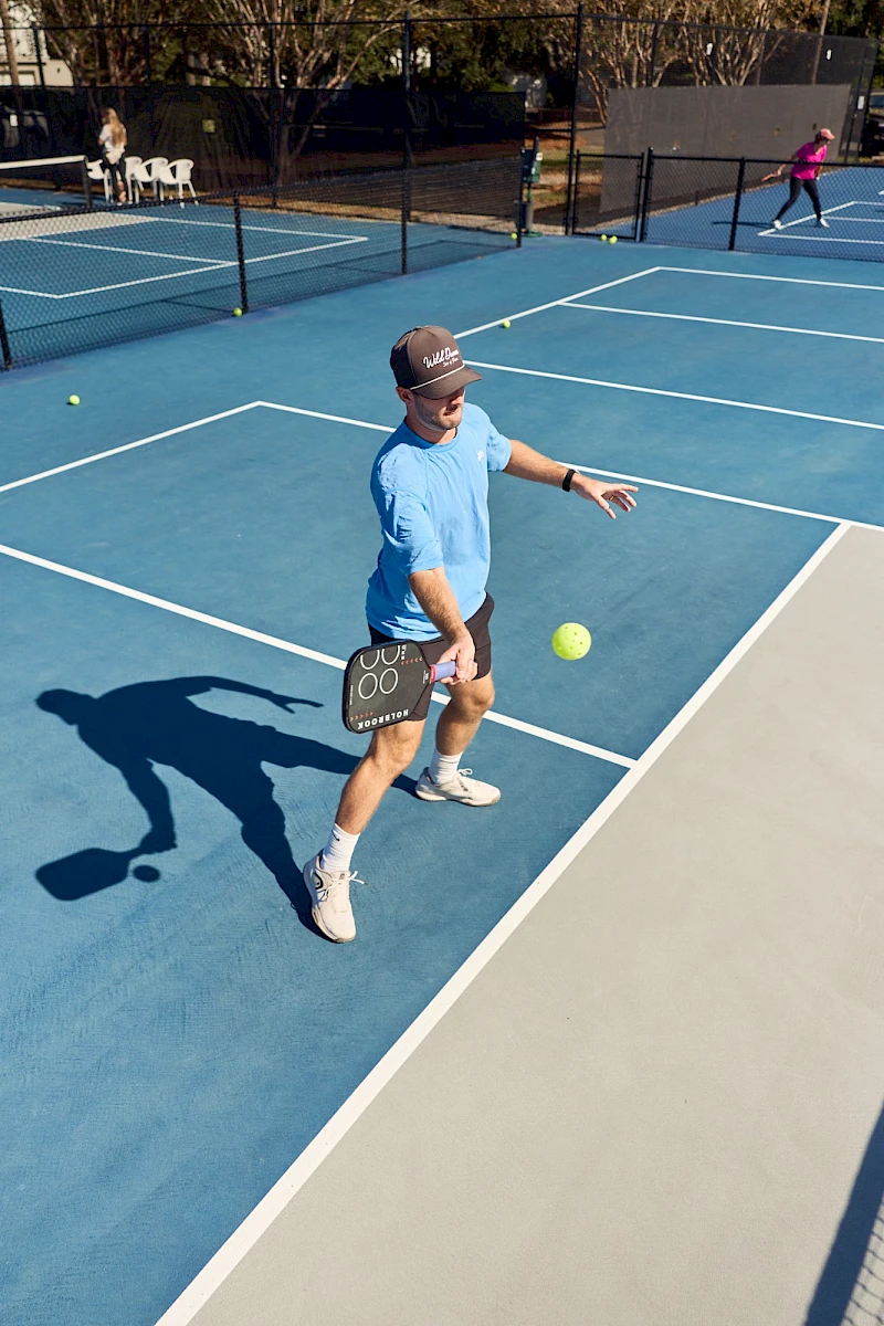 A man in a light blue shirt and white shorts throws a padel racket toward a tennis ball on a blue court, shadow extending beside him.
