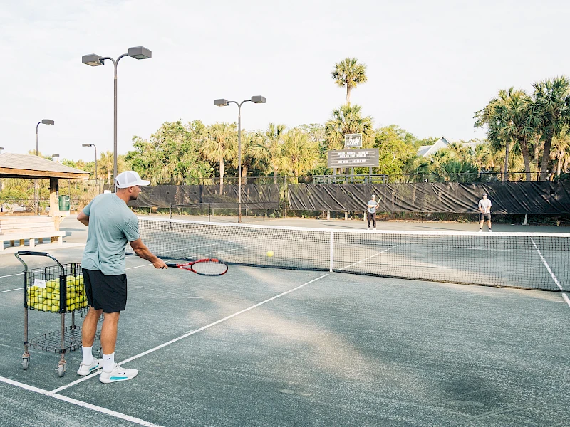 An older man practices tennis on a sunny outdoor court with a ball cart nearby and a few players in the background.