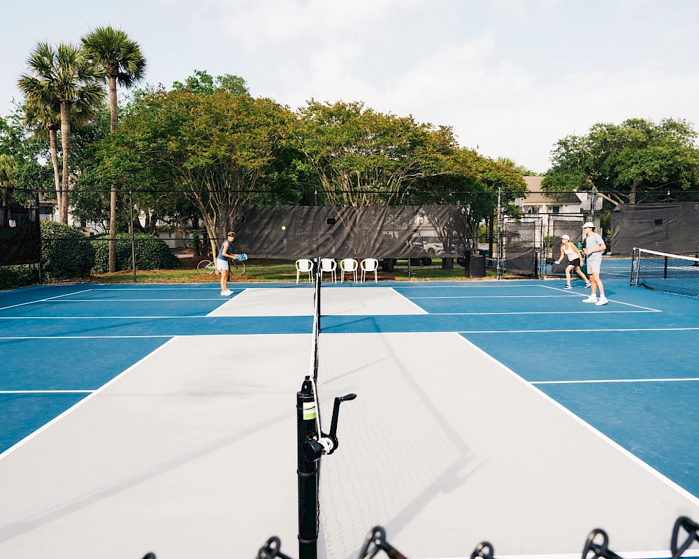 A beach volley-court with blue and white playing surface, net in center, and people in the background near the net.