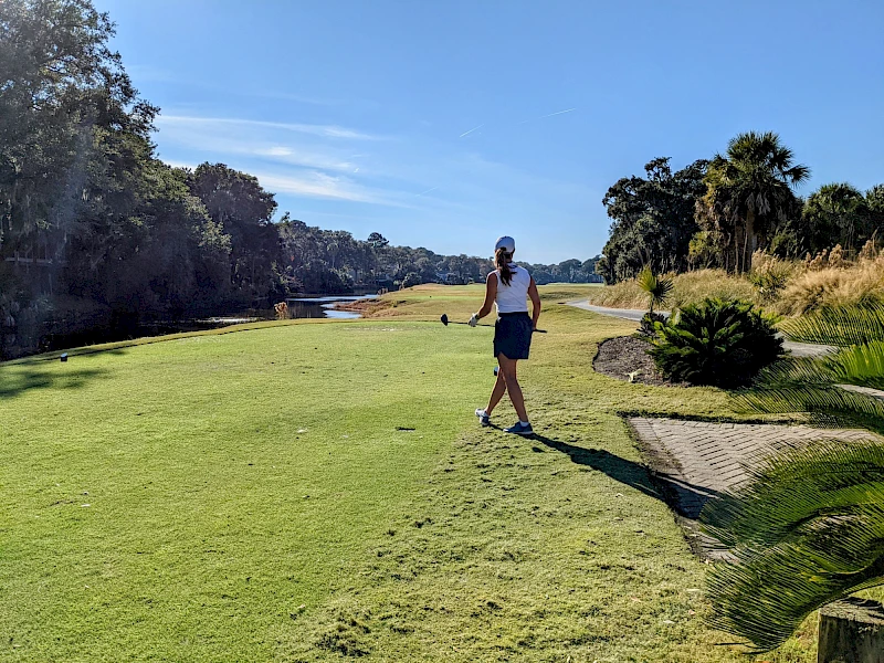 A person stands on a grassy golf fairway near a path, with trees and blue sky in a sunny, open park-like setting.