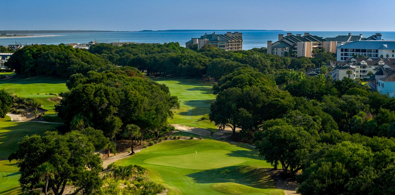 A coastal golf course with green fairways, trees, and buildings near the shoreline in the background.