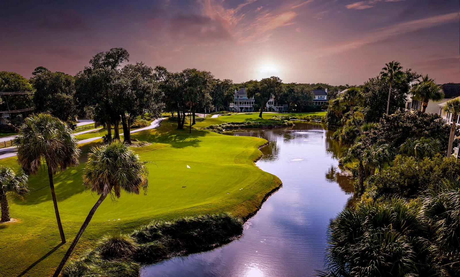 A scenic golf course at sunset: lush green fairways, a winding waterway, palm trees, and a calm reflection of the sky in the water, serene and inviting.