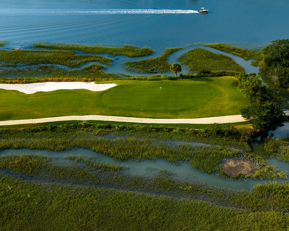 Aerial view of a golf green surrounded by water channels and marsh, with a white path and a boat visible in the blue water.